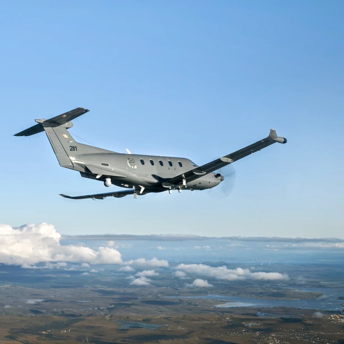 A Pilatus PC-12 aircraft in military livery flies over a landscape with clouds.