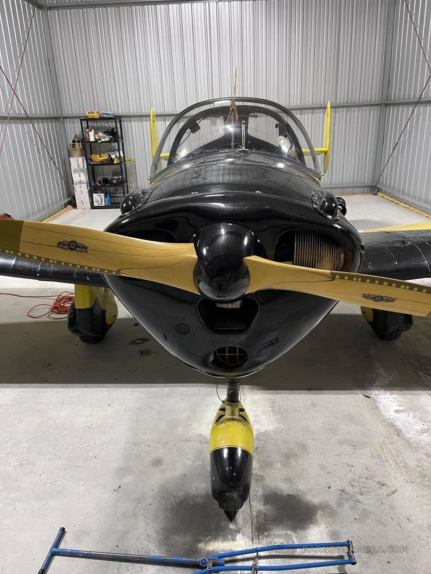A head-on view of a black and yellow single-engine aircraft with a wooden propeller, parked inside a metal hangar.