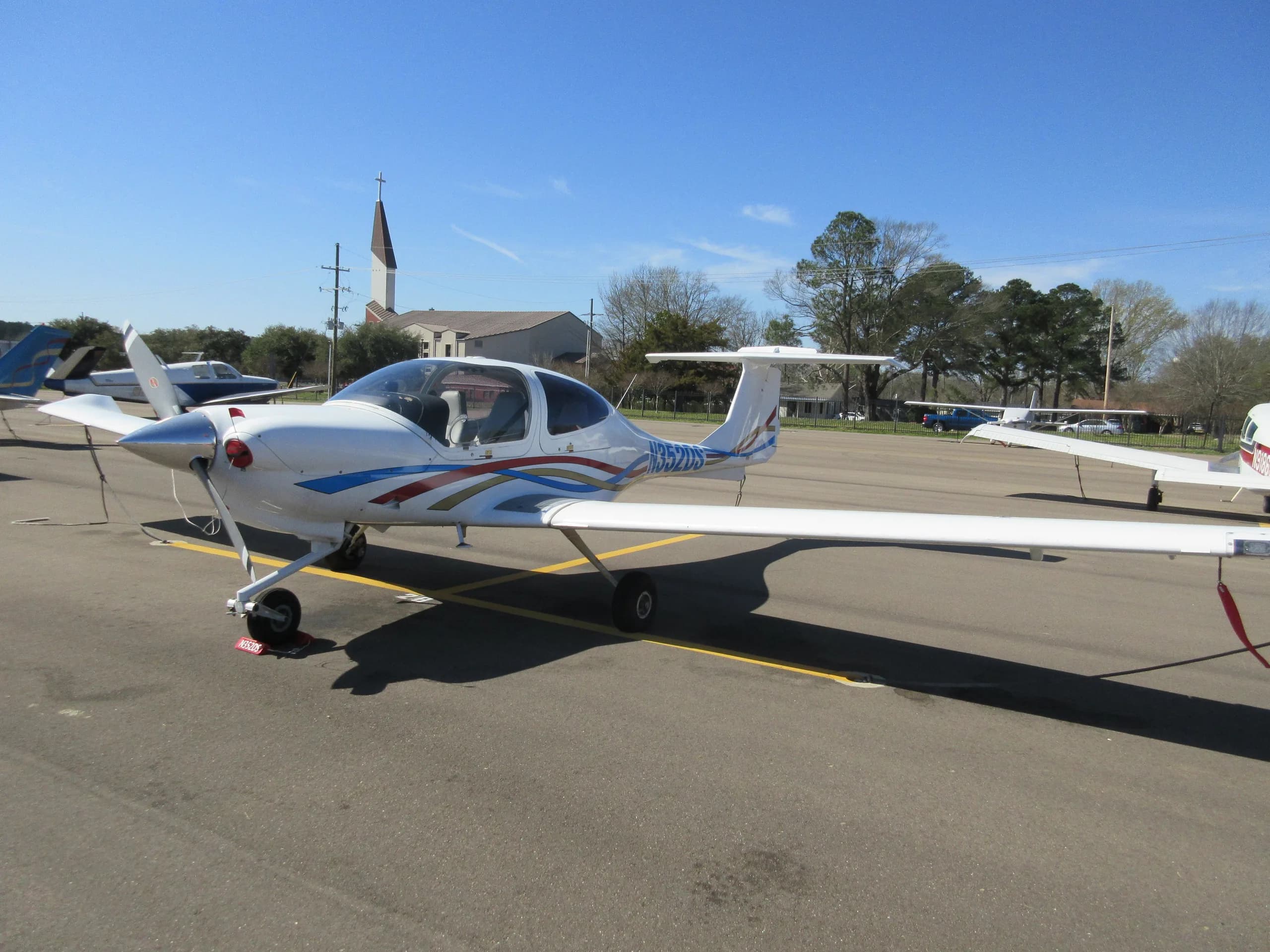 A white Diamond DA40 aircraft with blue, red, and gold stripes, registration N352DS, parked on an airport tarmac, viewed from the front-left.