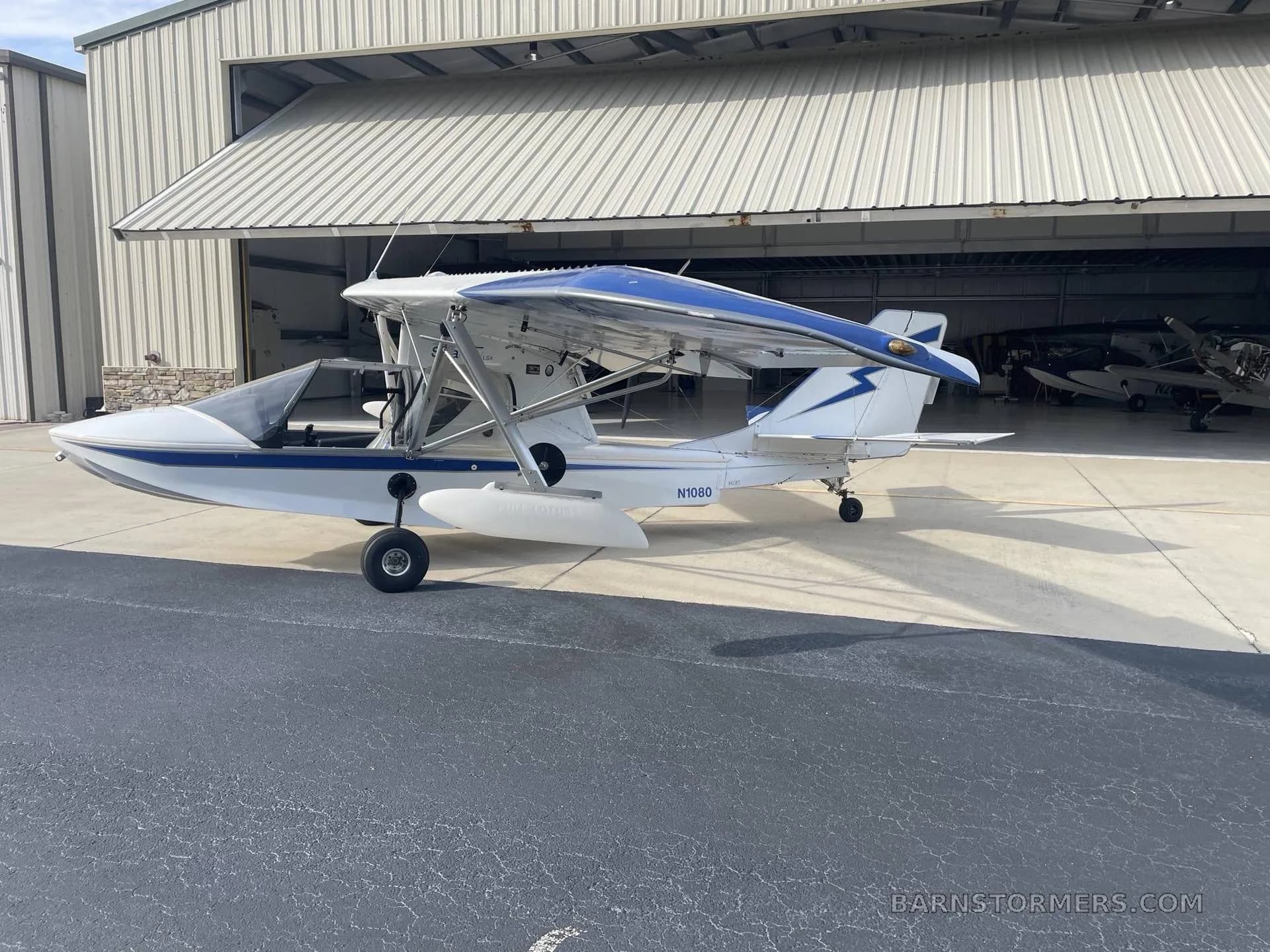 A white and blue Searey amphibious aircraft (N1080) parked on an airfield in front of an open hangar.