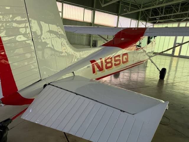 A rear quarter view of the red and white Cessna 180/185 aircraft, tail number N8SQ, in a hangar.