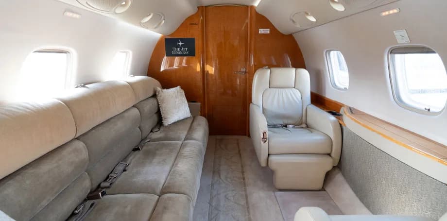 The rear section of a private jet cabin, featuring a long beige sofa and a single light leather armchair.