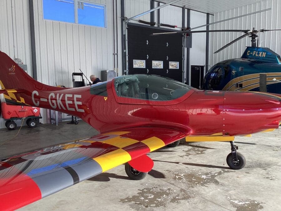Side view of a red VL3 Evolution aircraft parked inside a hangar next to a helicopter.
