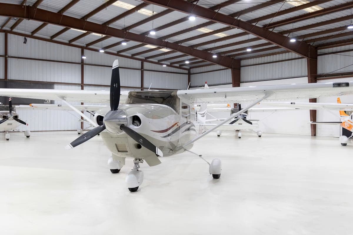 A front view of a white and grey Cessna Stationair TC (N6045B) with a black propeller, parked in a hangar.