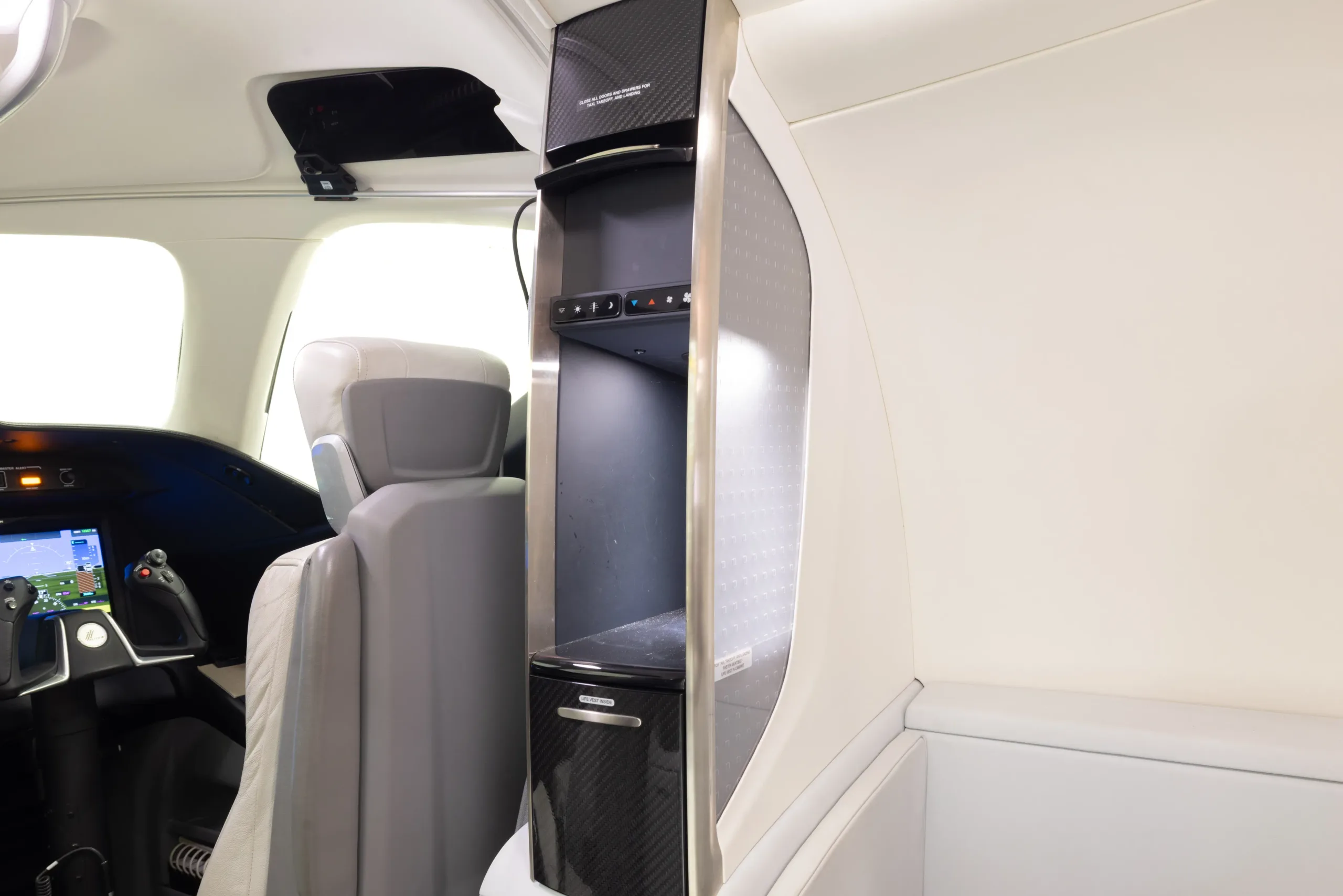 A view inside the aircraft cabin showing a galley unit and the back of a pilot's seat, with a glimpse of the cockpit.
