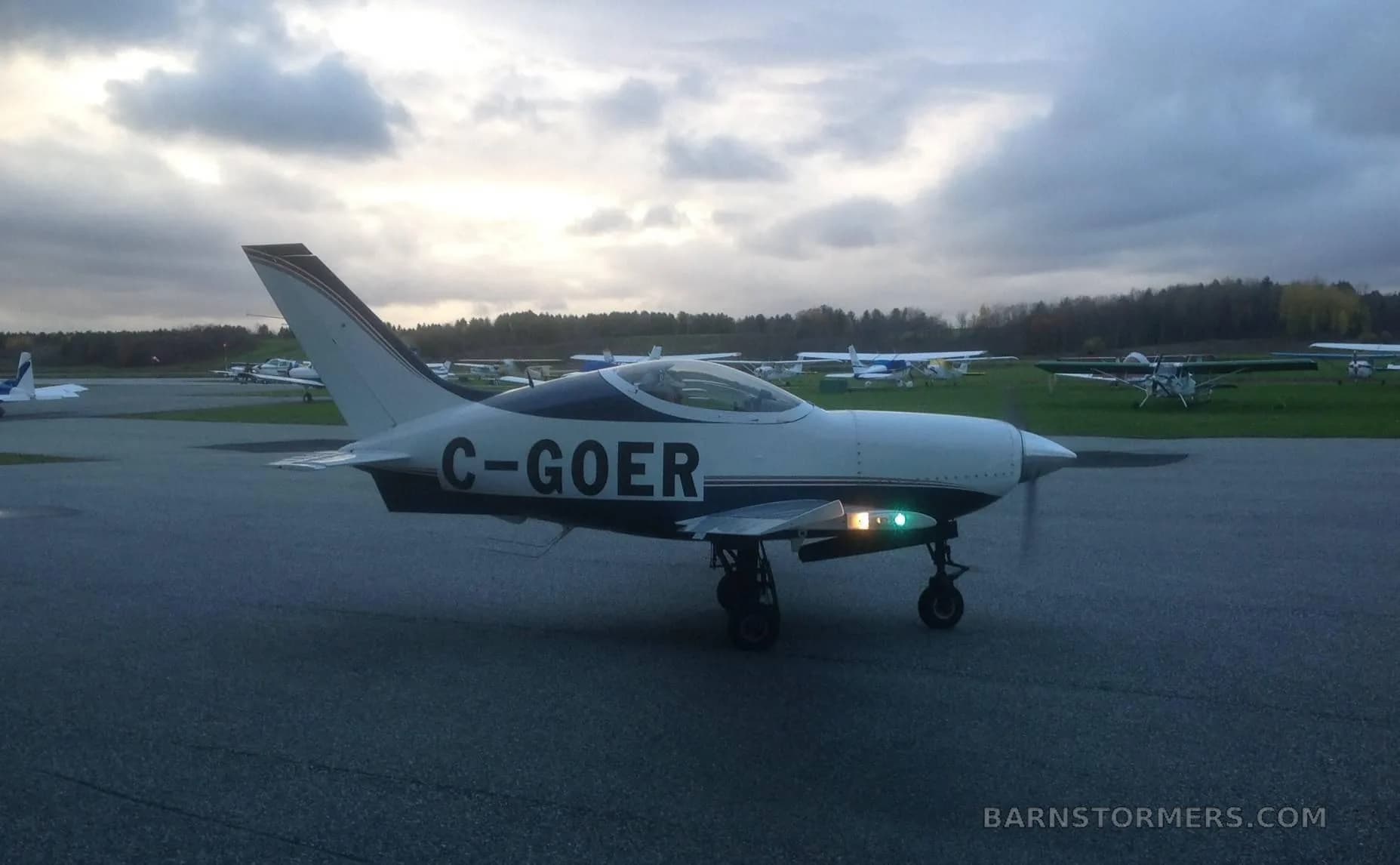 A white and blue single-engine aircraft with its landing lights on, taxiing on an airfield under a cloudy sky.