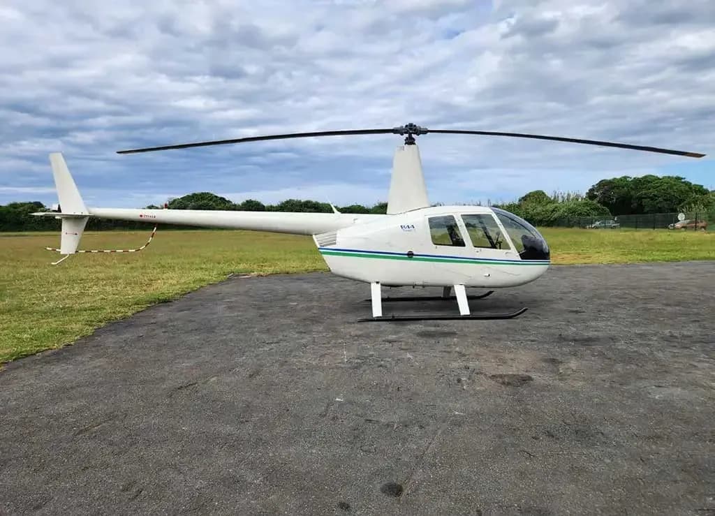 A white Robinson R44 helicopter with green and blue stripes, viewed from the side, parked on an asphalt pad.