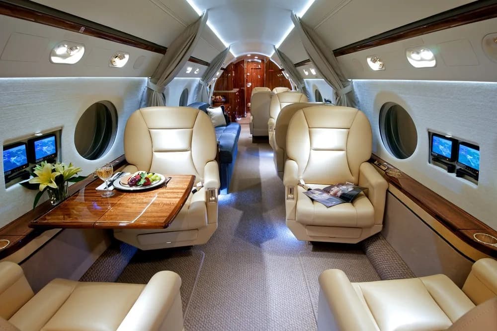 The luxurious cabin interior of a private jet, featuring tan leather seats, a wooden table with a meal, and individual entertainment screens.
