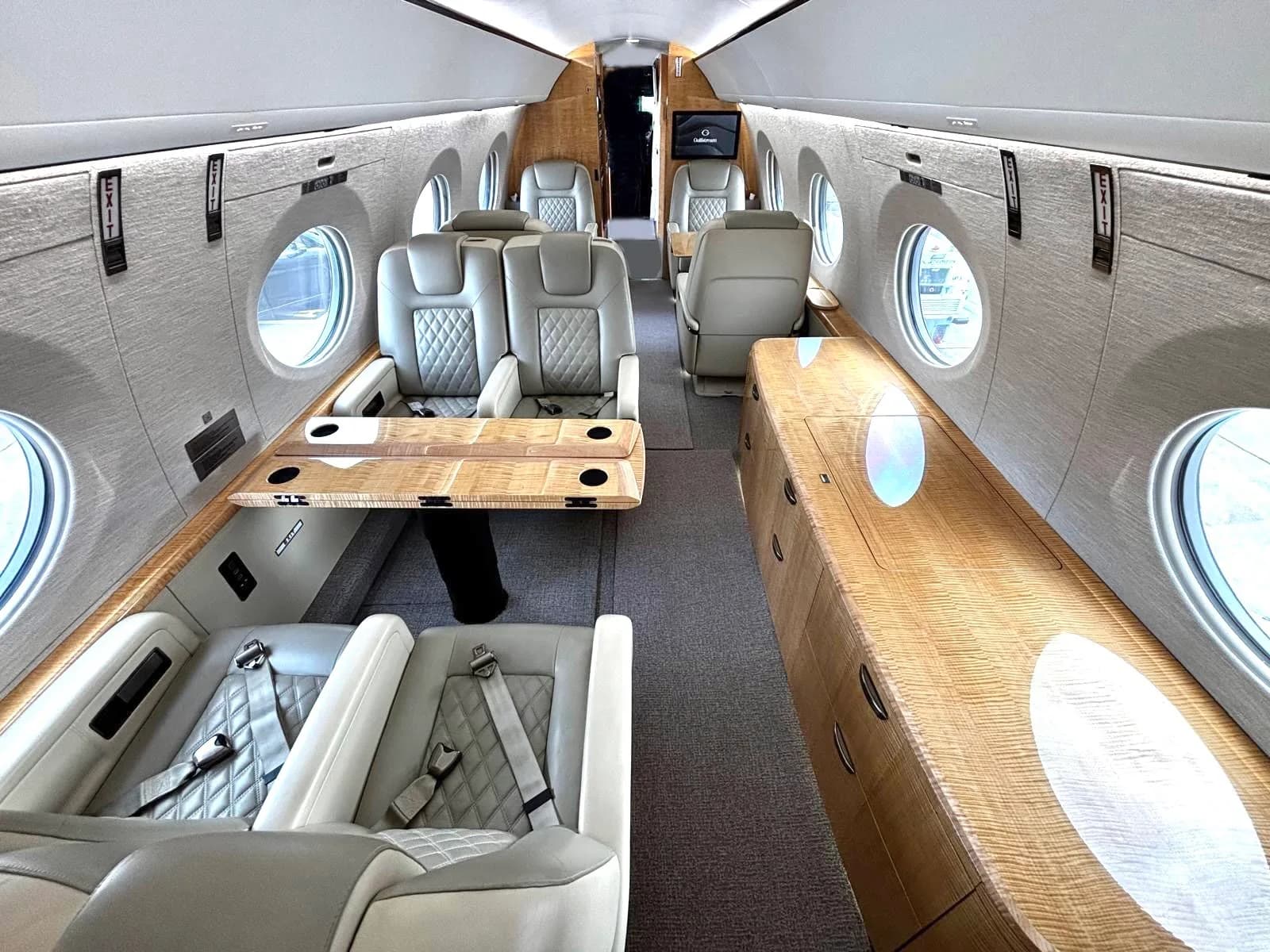 A view down the aisle of a private jet cabin, showing light-colored quilted seats, a fold-out table, and wooden cabinetry.