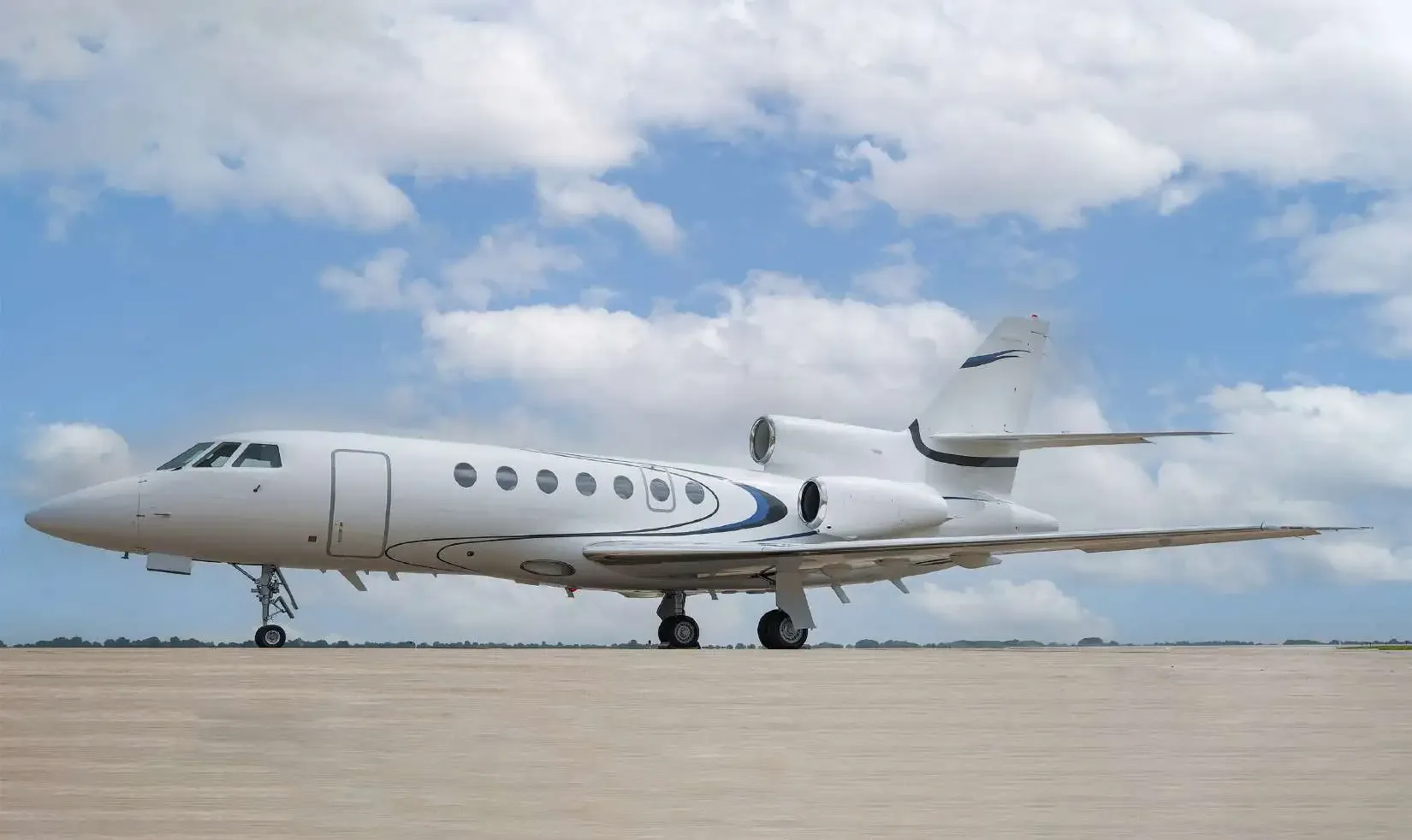 A white Dassault Falcon business jet with blue and black stripes on the tail and engines, parked on an airfield under a cloudy sky.