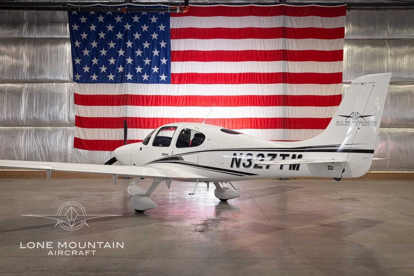Side view of a white Cirrus SR22 aircraft with black stripes and registration N327TM, parked in a hangar with an American flag backdrop, with its gull-wing doors closed.