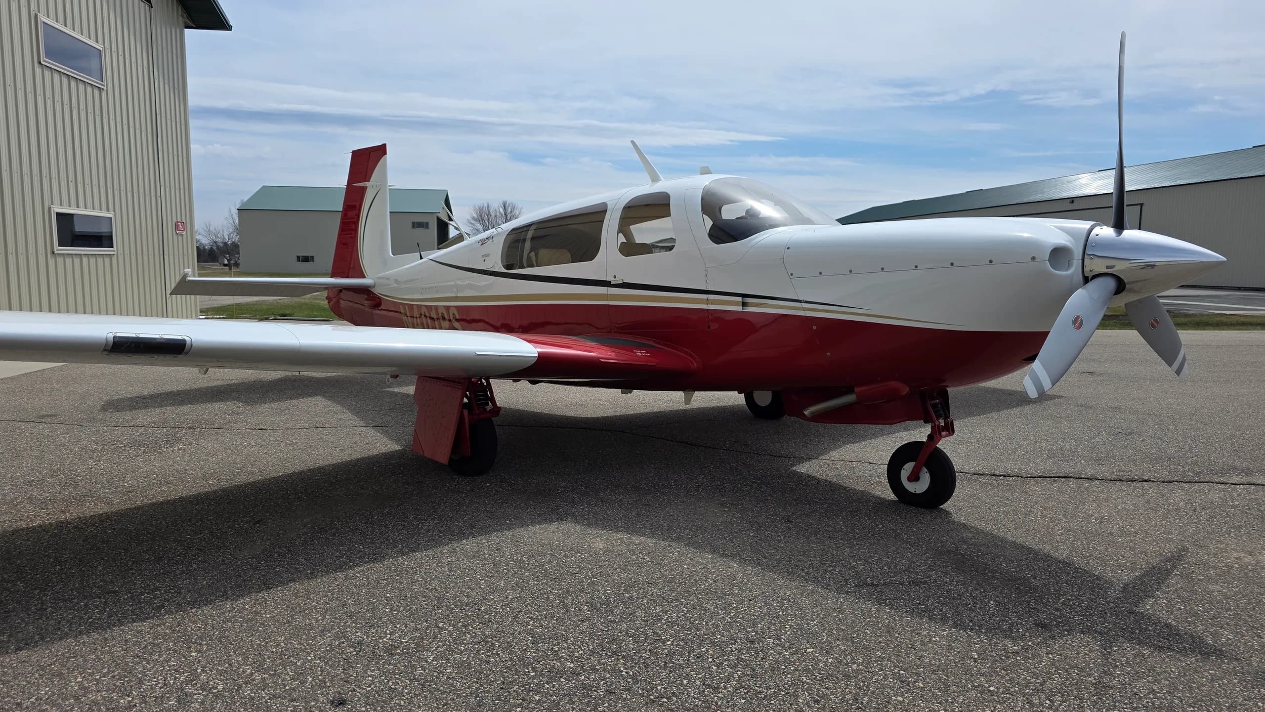 A red and white Mooney aircraft with gold stripes parked on an asphalt apron, viewed from the front right.