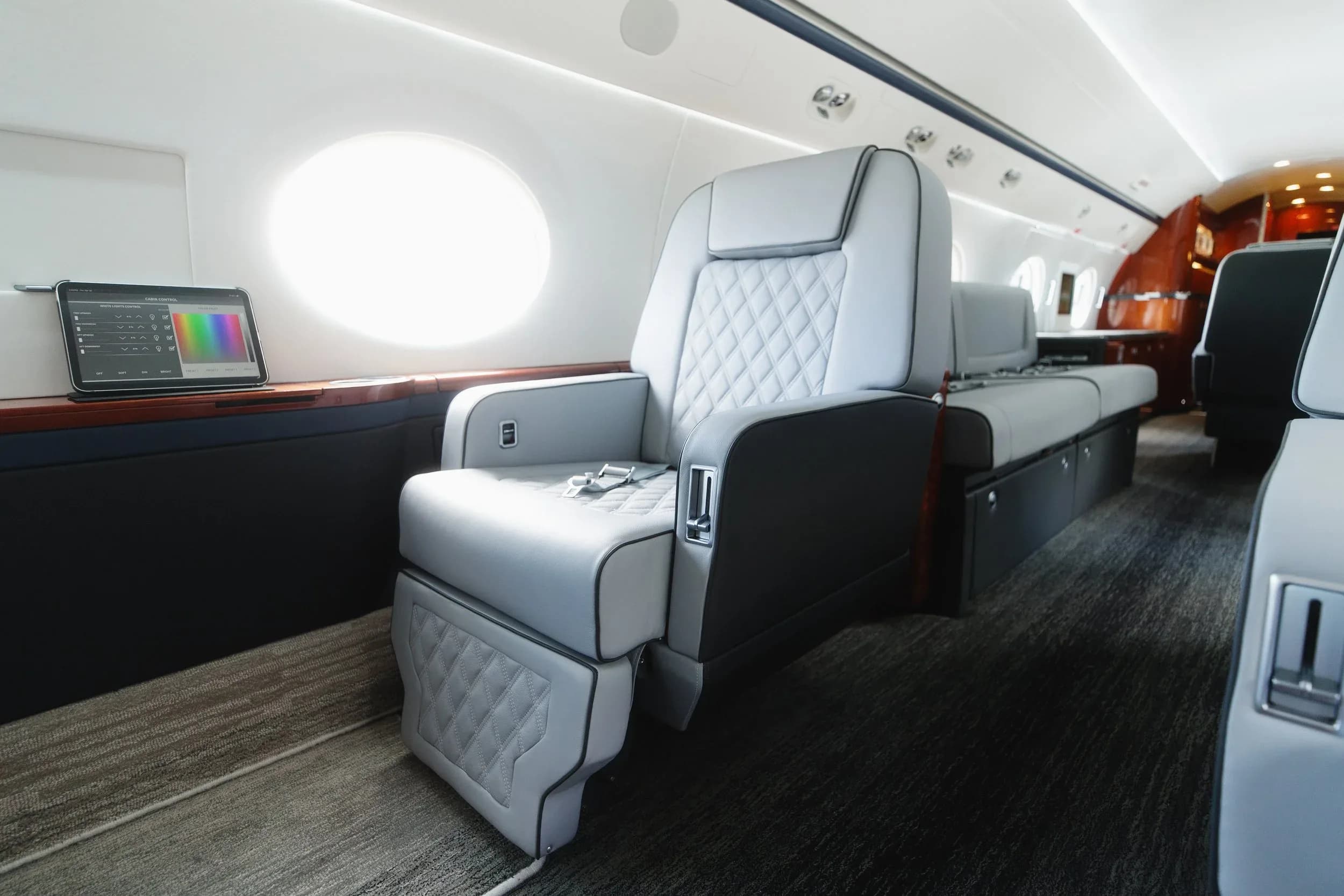 A close-up view of a light gray quilted leather seat in the private jet's cabin, with a tablet on the side table.