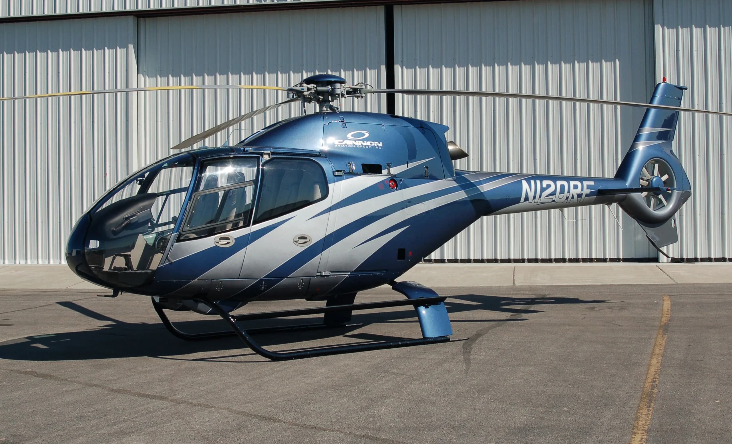 A blue and silver Eurocopter EC120 Colibri helicopter parked in front of a metal hangar.