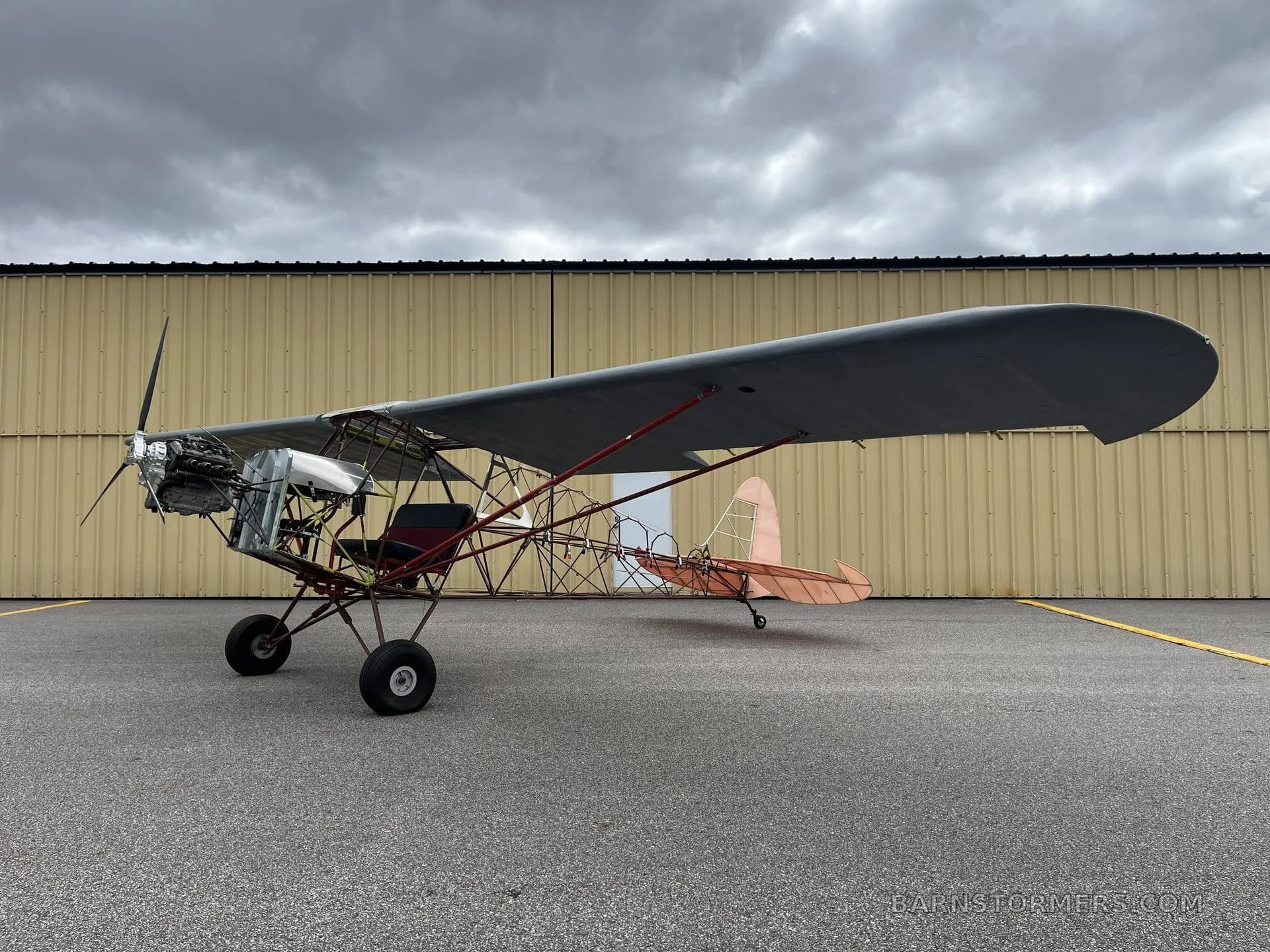 A side view of an experimental ultralight aircraft with an exposed frame, a single wing, and a propeller, parked on an asphalt apron in front of a yellow hangar.