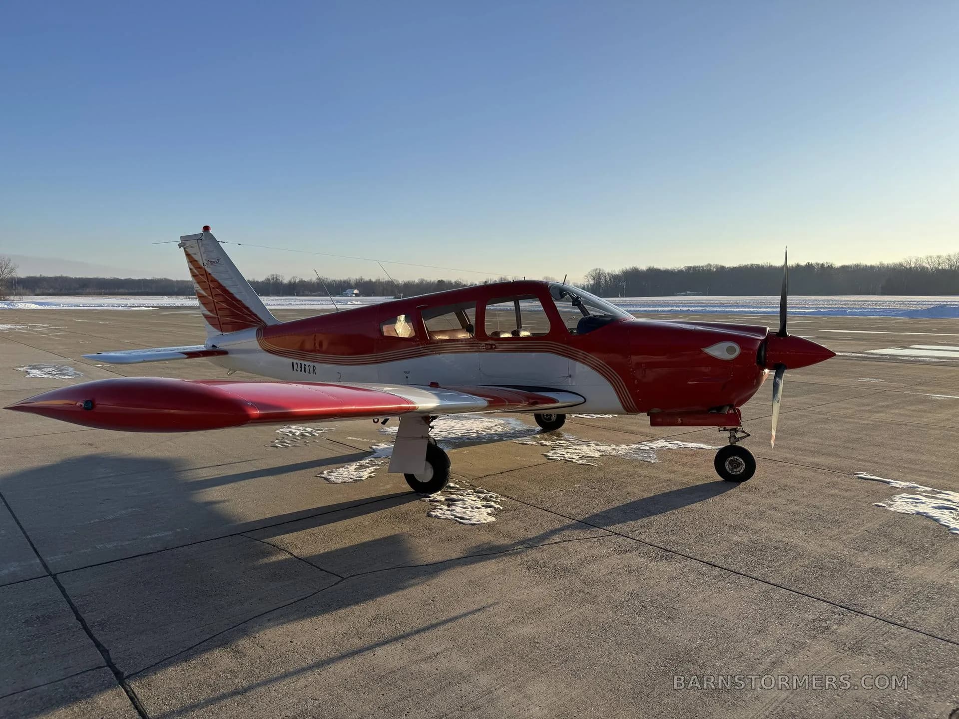 A red and white Mooney M20 series aircraft parked on a snowy tarmac under a clear blue sky.