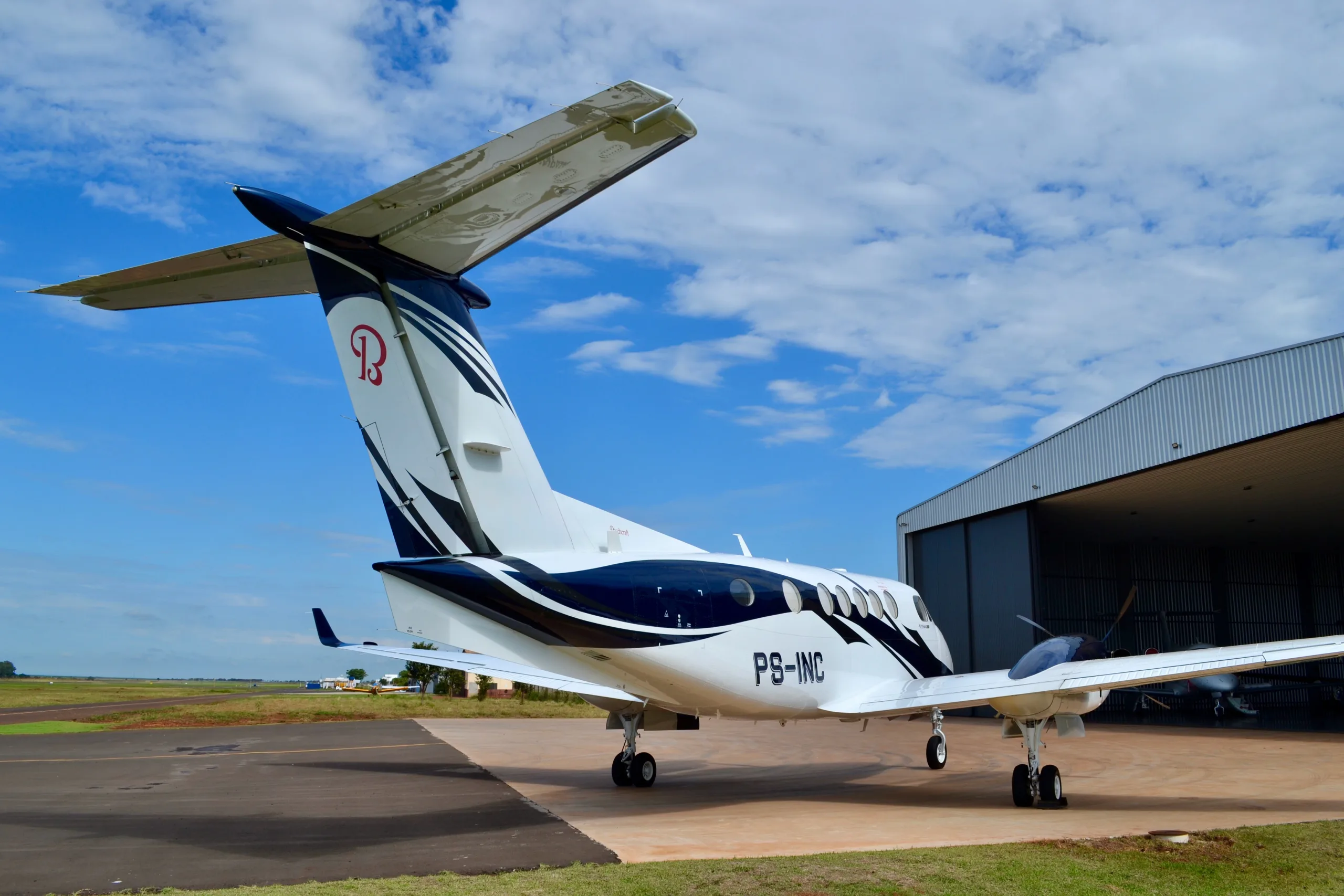 A rear-side view of a white and blue twin-turboprop aircraft, registration PS-INC, parked on a tarmac near a hangar.