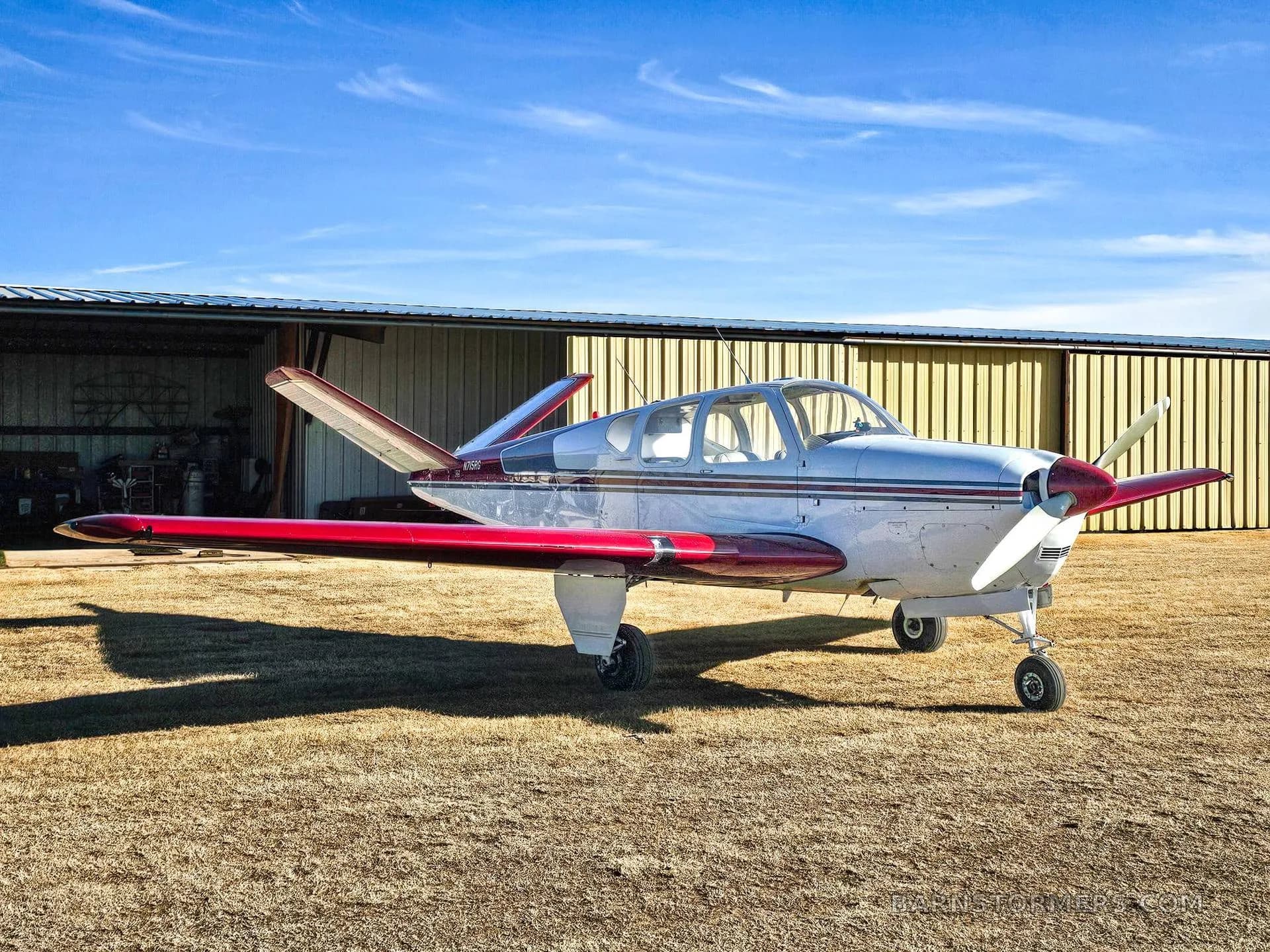 A white and red Mooney M20 series aircraft parked on a grassy field in front of an open hangar under a blue sky.