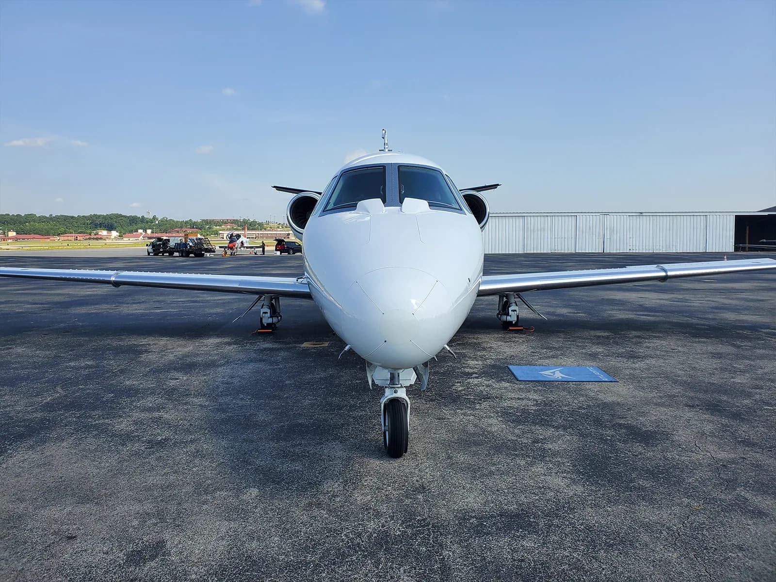 A head-on view of a white private jet with gold and black stripes, parked on an asphalt tarmac with other aircraft in the background.