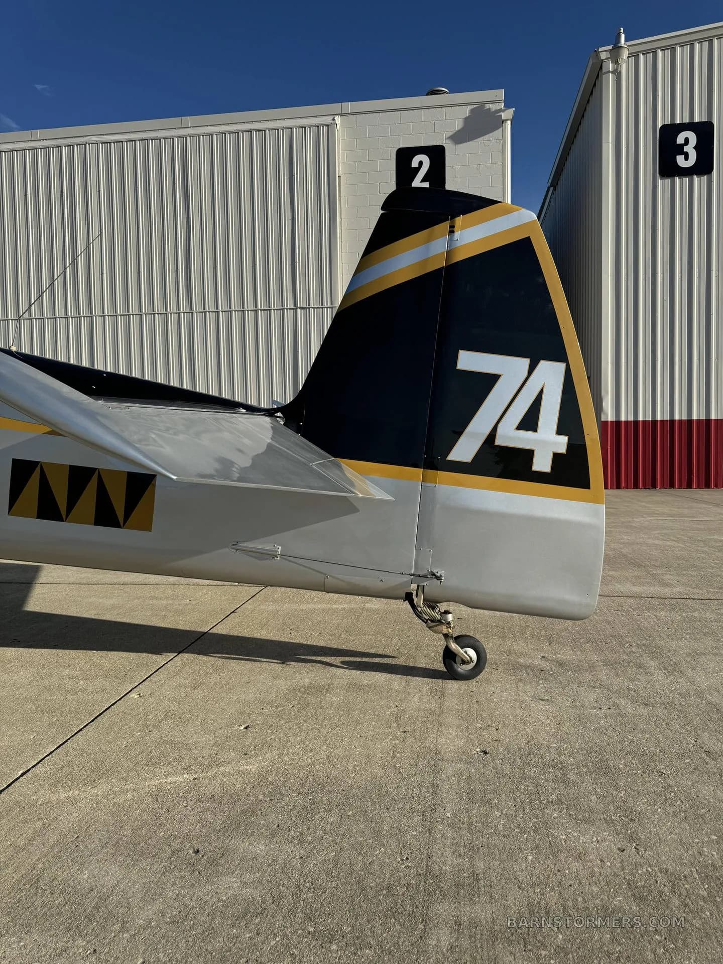 The tail section of a silver aircraft with black and gold stripes and the number "74" is shown outdoors next to hangars.