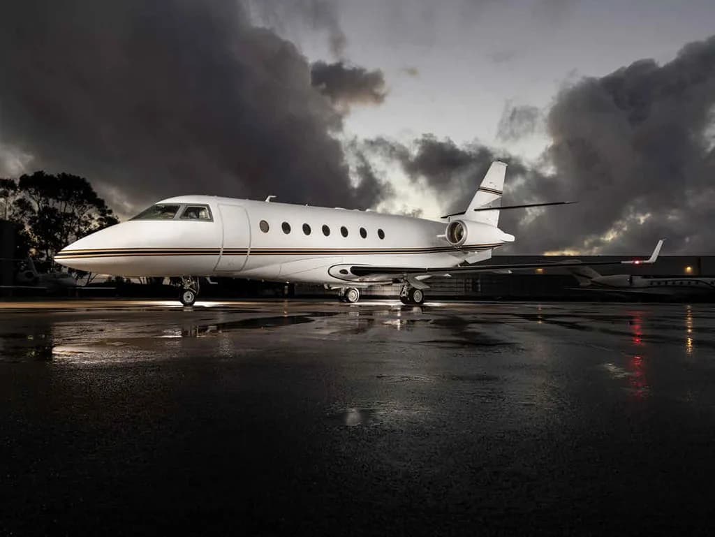 A white Gulfstream G450 business jet with a gold stripe on a wet tarmac at dusk.