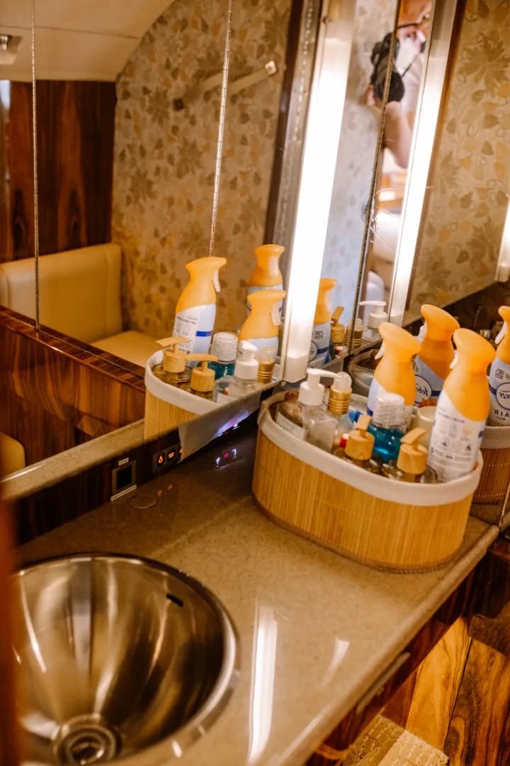 An aircraft lavatory with a stainless steel sink, a mirror, and various toiletries on the counter.