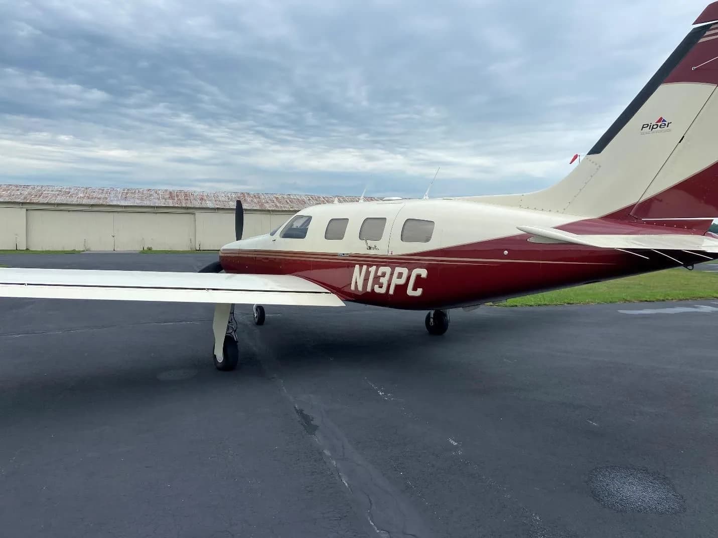 Side view of a Piper Malibu Mirage (N13PC) in maroon and cream paint scheme, parked on an asphalt ramp under a cloudy sky.