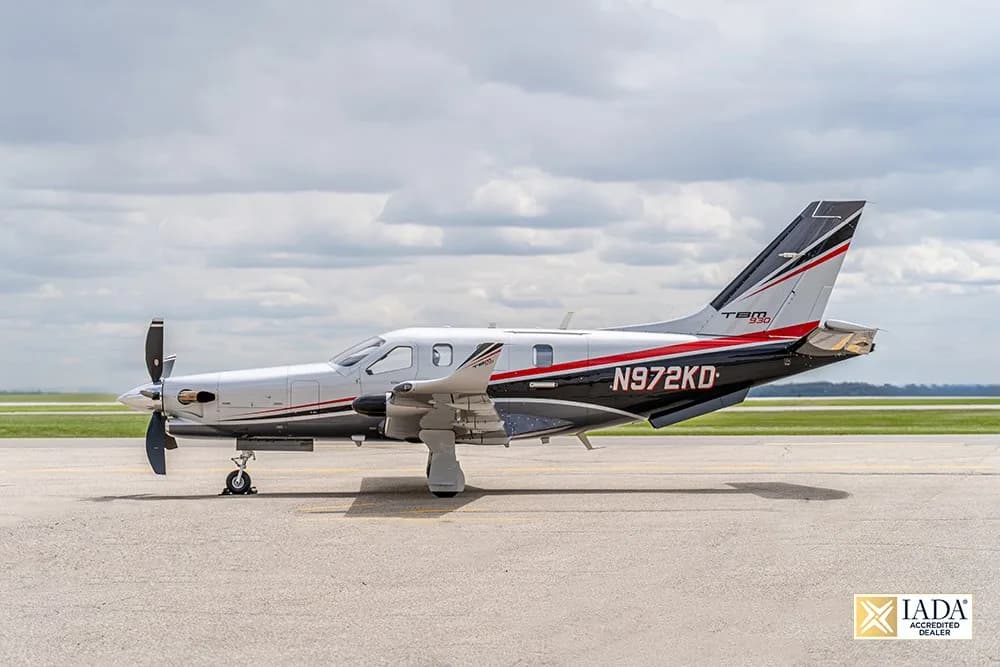 A side view of a silver and black Daher TBM 930 aircraft with red accents, registration N972KD, on an airfield under a cloudy sky.