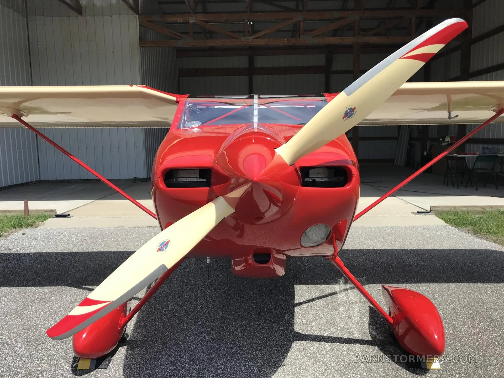 A front view of a red and cream single-engine aircraft with a large propeller, parked inside a hangar.