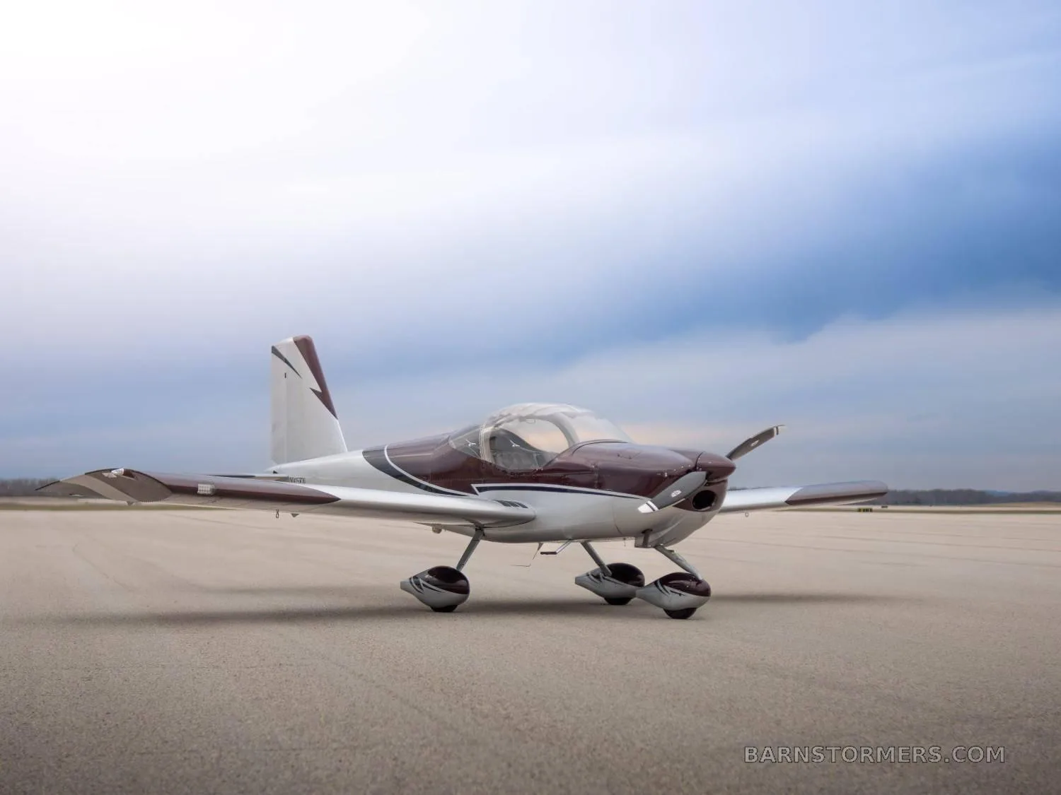 A low-angle front-side view of a two-seat, single-engine propeller aircraft with a maroon and silver paint scheme on an airfield under a cloudy sky.