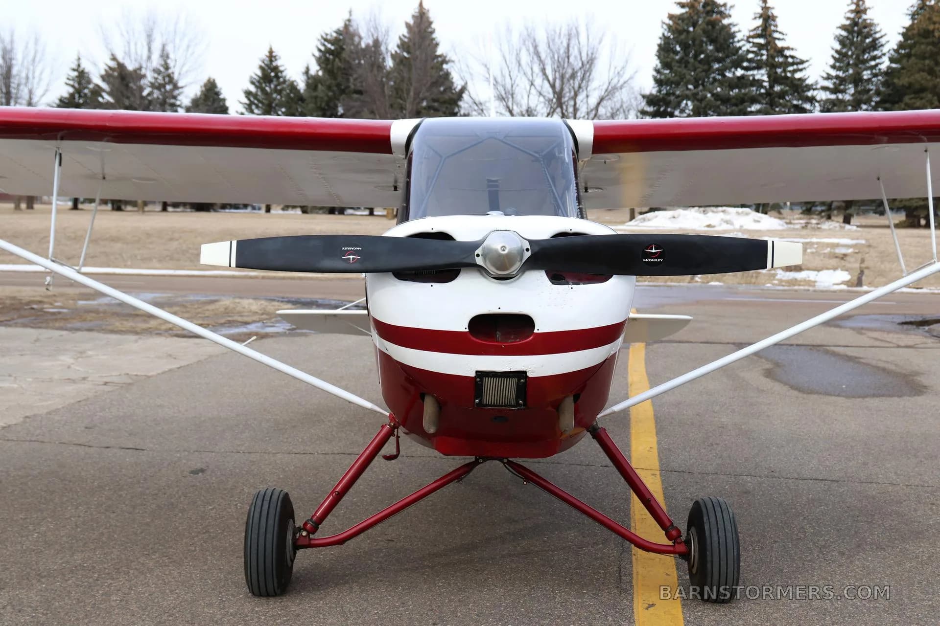 A head-on view of the red and white N4715E taildragger aircraft, highlighting its propeller and front landing gear.