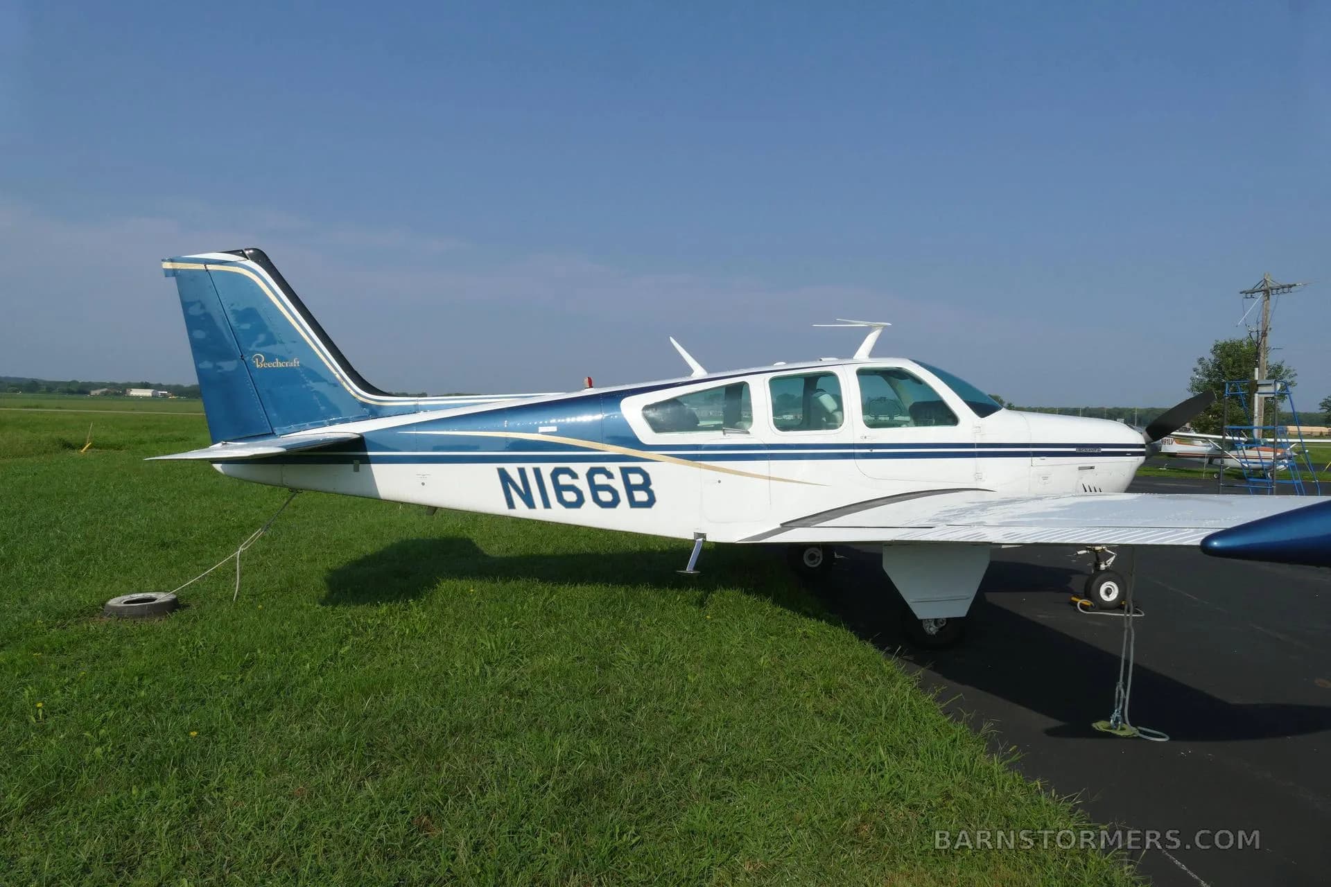 A side view of a white and blue Beechcraft Bonanza (N166B) parked on the edge of an airfield tarmac.