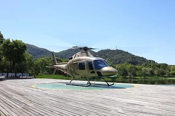A tan and camouflage-painted helicopter rests on a wooden helipad next to a lake with green mountains in the background.