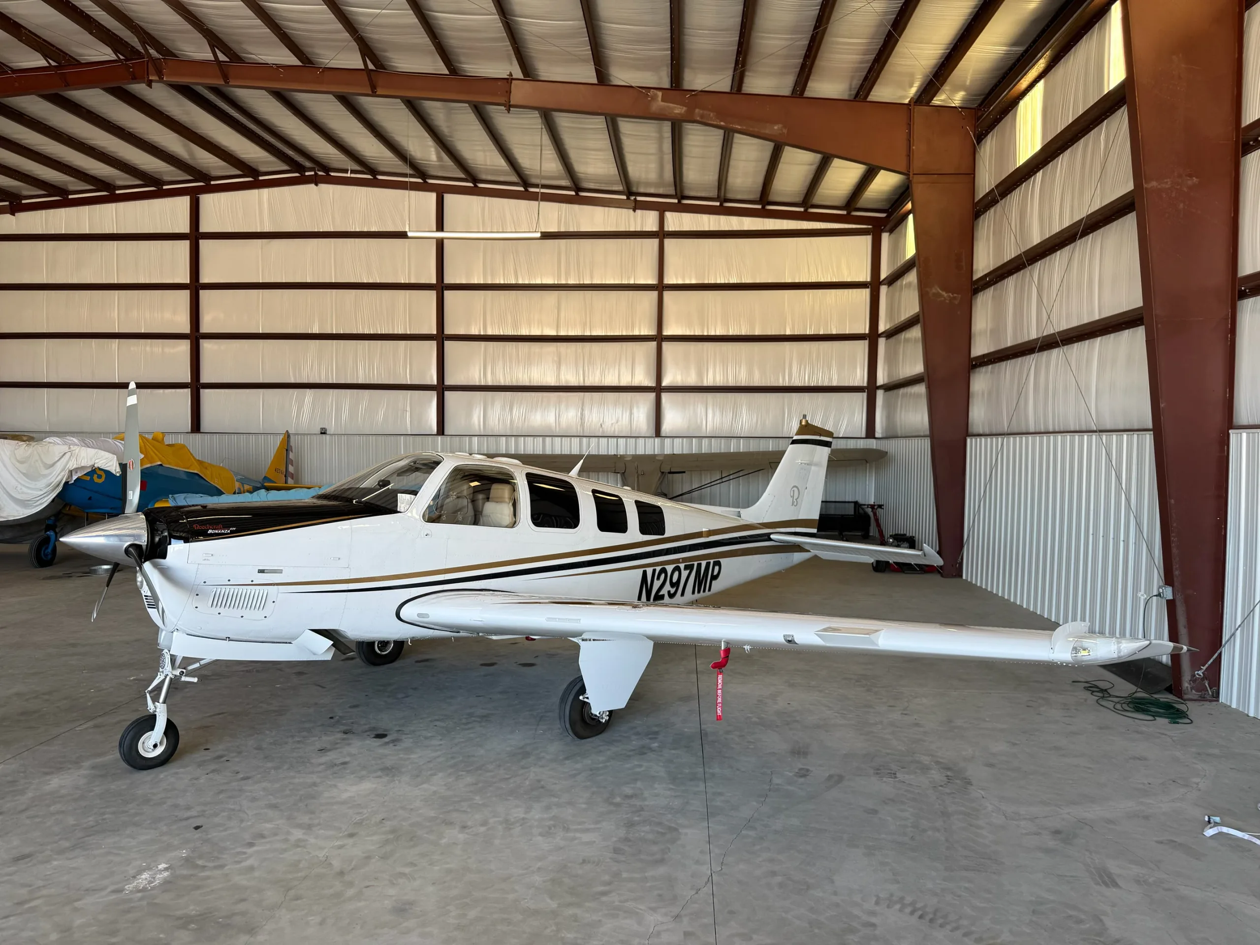 A white Beechcraft Bonanza N297MP with black and gold stripes parked inside a hangar.