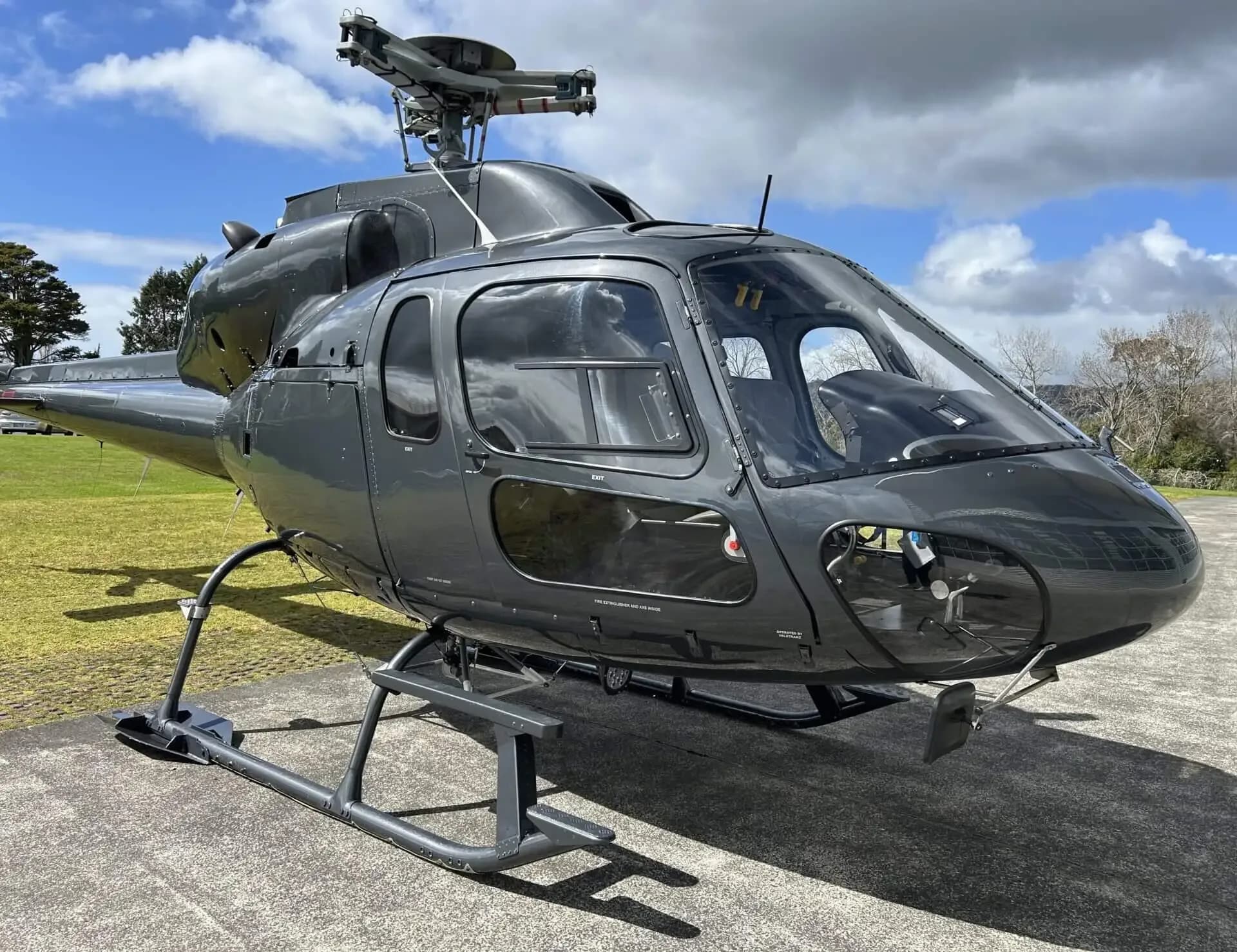 A dark grey Eurocopter AS355 F1 helicopter viewed from the front-left, parked on a concrete pad under a bright sky.