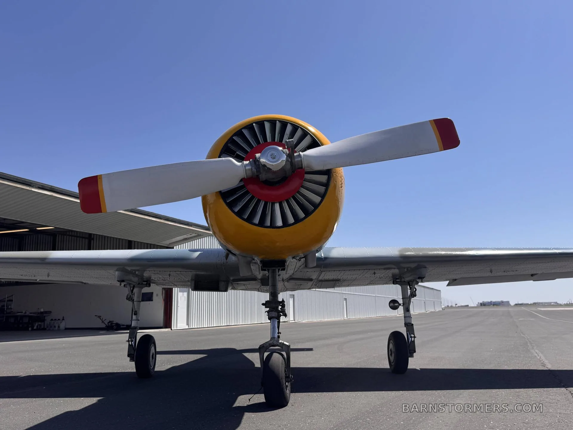 A front view of a Yak-502GC aircraft with a yellow nose and a large propeller, parked on an asphalt tarmac in front of hangars.