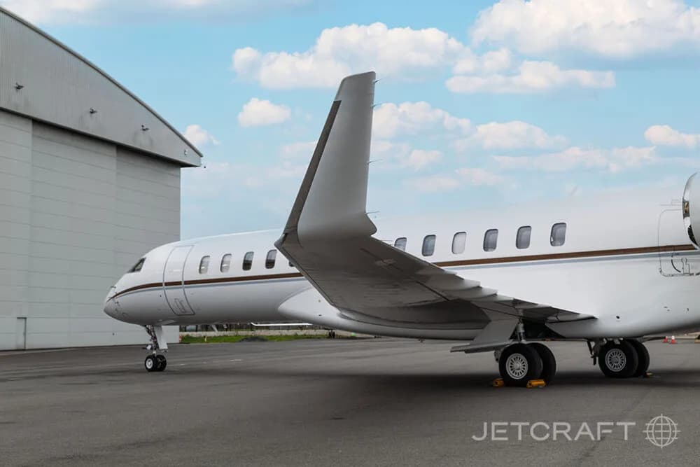 The rear fuselage and tail of a white Bombardier Global business jet parked next to a large hangar.