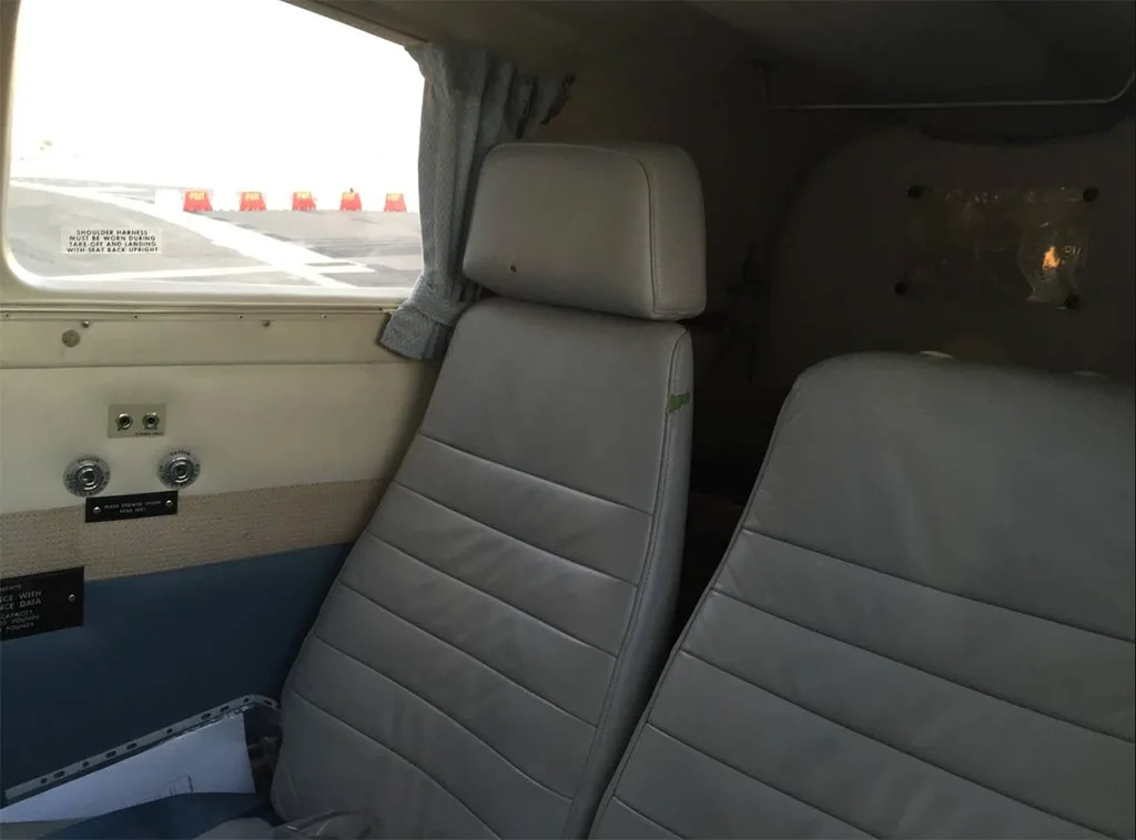 Interior view of an aircraft cabin showing two light gray leather seats and a window looking out onto an airfield.