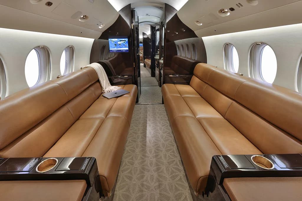 The spacious interior of a business jet shows two long tan leather sofas facing each other, with a patterned carpeted aisle.