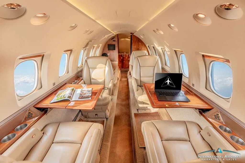 The luxurious interior cabin of a private jet features beige leather seats, wooden tables, and a laptop on one table.