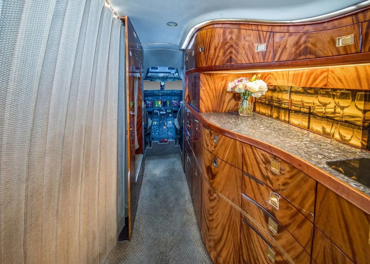 A view down the aircraft aisle from the galley, featuring polished wooden cabinets, a granite countertop with flowers, and the cockpit visible in the distance.