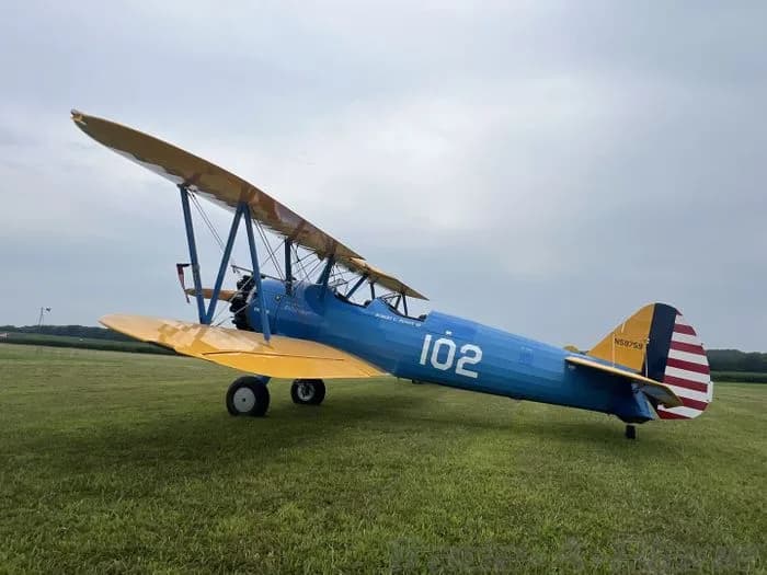 A blue and yellow Boeing Stearman biplane is parked on a grassy field.