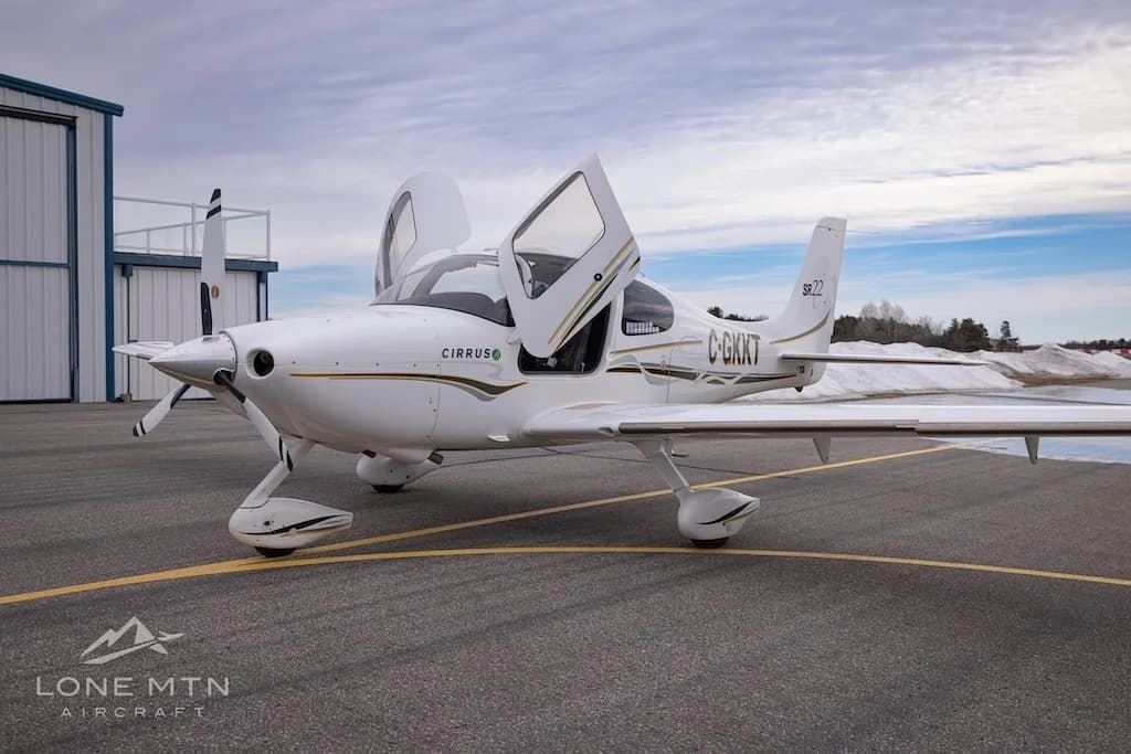 A white Cirrus SR22 aircraft with registration C-GKKT parked on an airfield with its doors open, a hangar in the background.