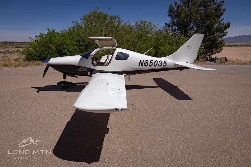 A white and black single-engine aircraft, N65035, with its gull-wing doors open, parked on an asphalt surface with trees and desert landscape behind it.