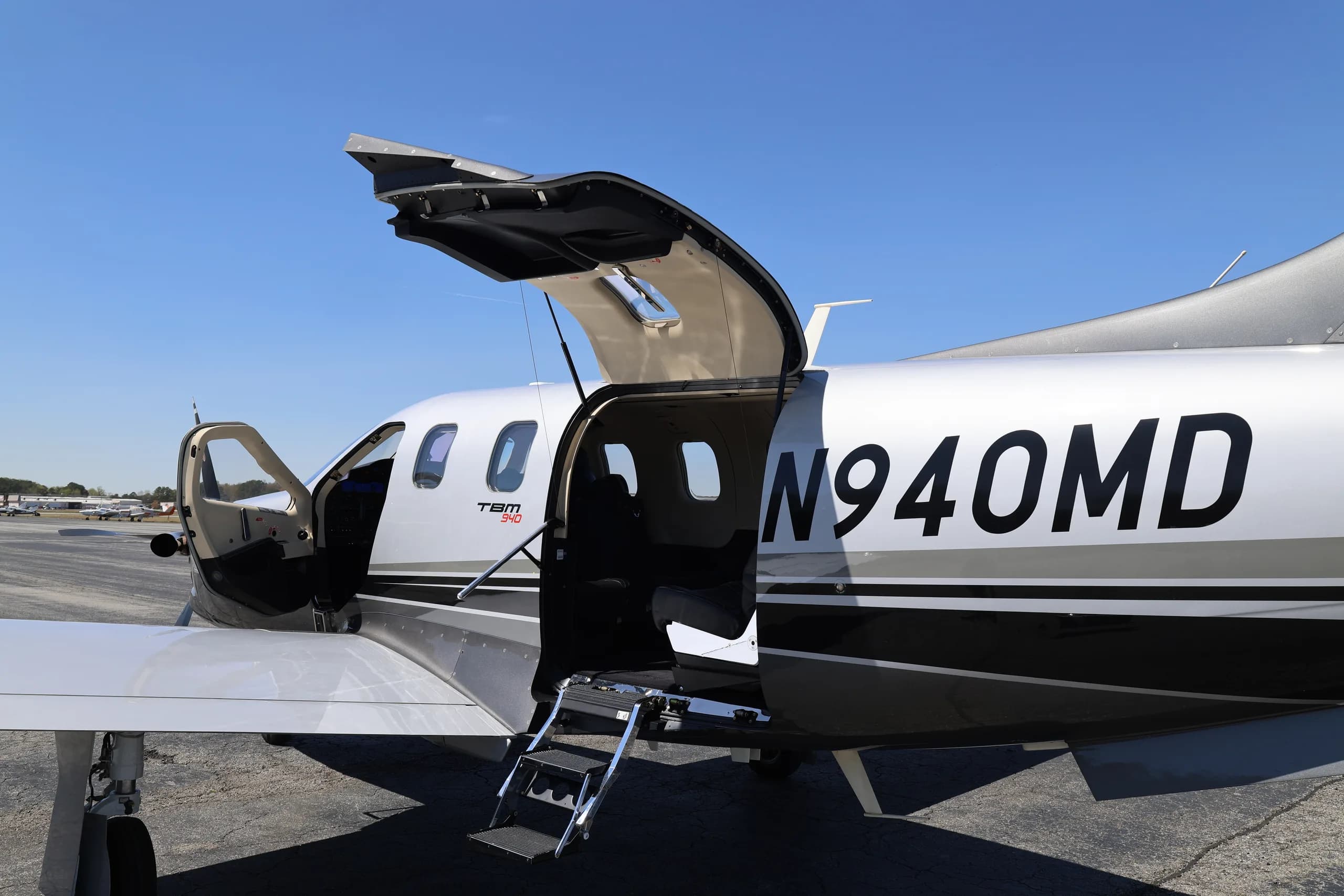 The side of a silver and black TBM 940 aircraft with registration N940MD, showing the open passenger and cargo doors with steps.