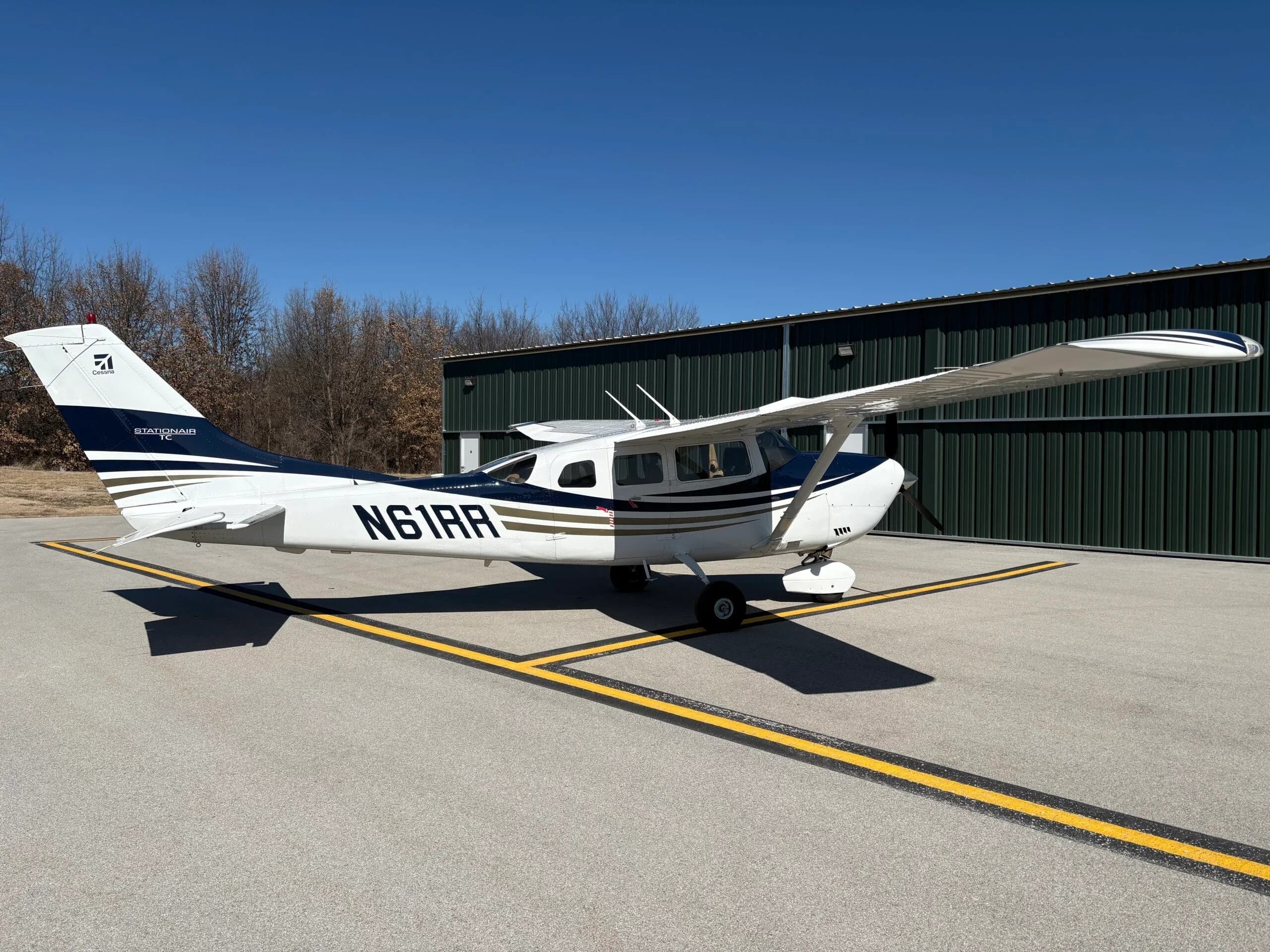 Three-quarter rear view of a white and dark blue Cessna Stationair TC aircraft with registration N61RR parked on an airfield.