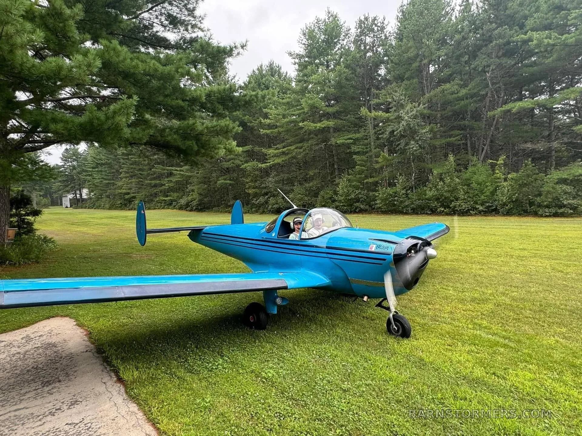 A bright blue Ercoupe aircraft with black stripes on a grassy field next to a paved path, with a pilot in the cockpit.