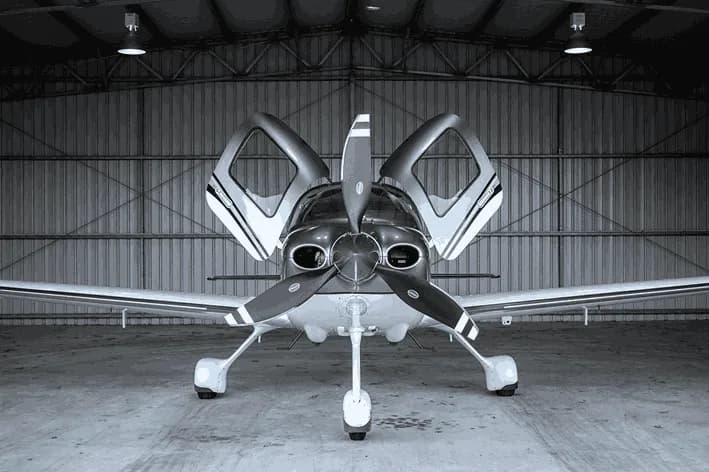 A light-colored propeller aircraft with open gull-wing doors is parked head-on inside a hangar.