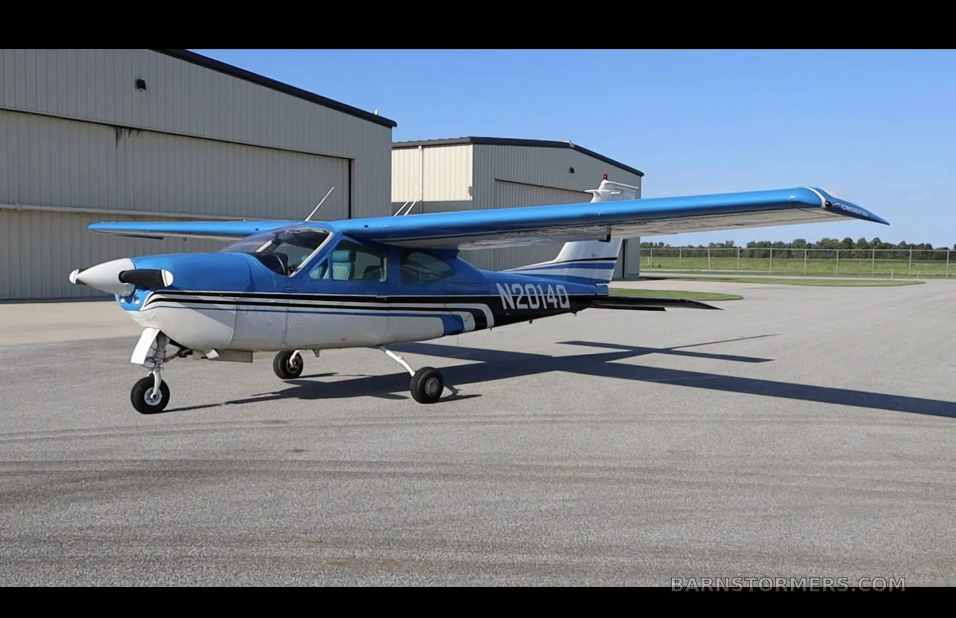 A blue and white Cessna 210 Centurion with registration N2014Q is parked on an asphalt ramp in front of hangars.