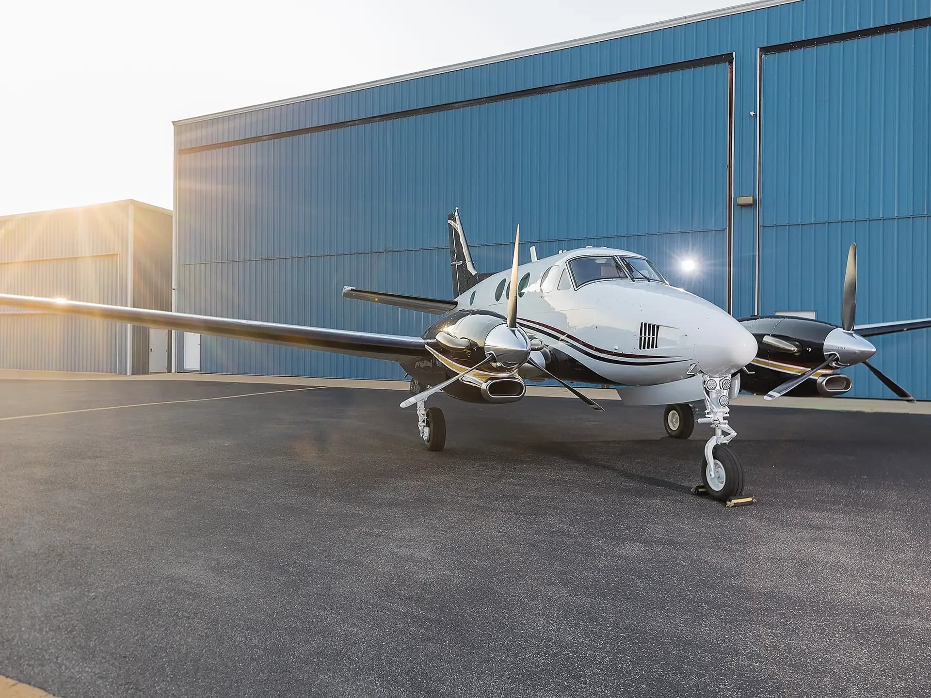 A white and black twin-engine turboprop aircraft with red stripes is parked on an asphalt ramp in front of blue hangars at sunset.